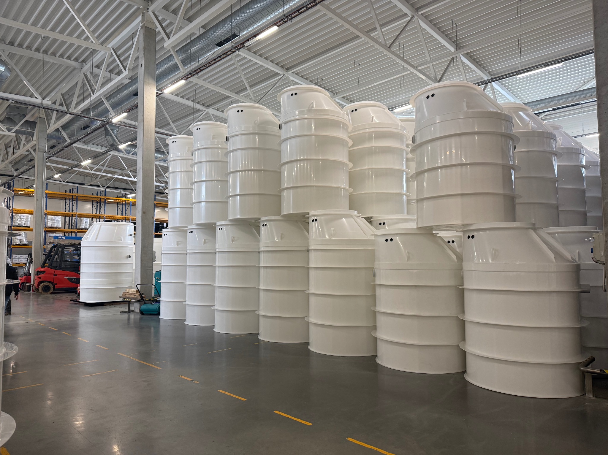 Magazyn pełen białych, plastikowych zbiorników na szambo lub oczyszczalnie ścieków, ustawionych w rzędach. W tle widoczny wózek widłowy i regały.