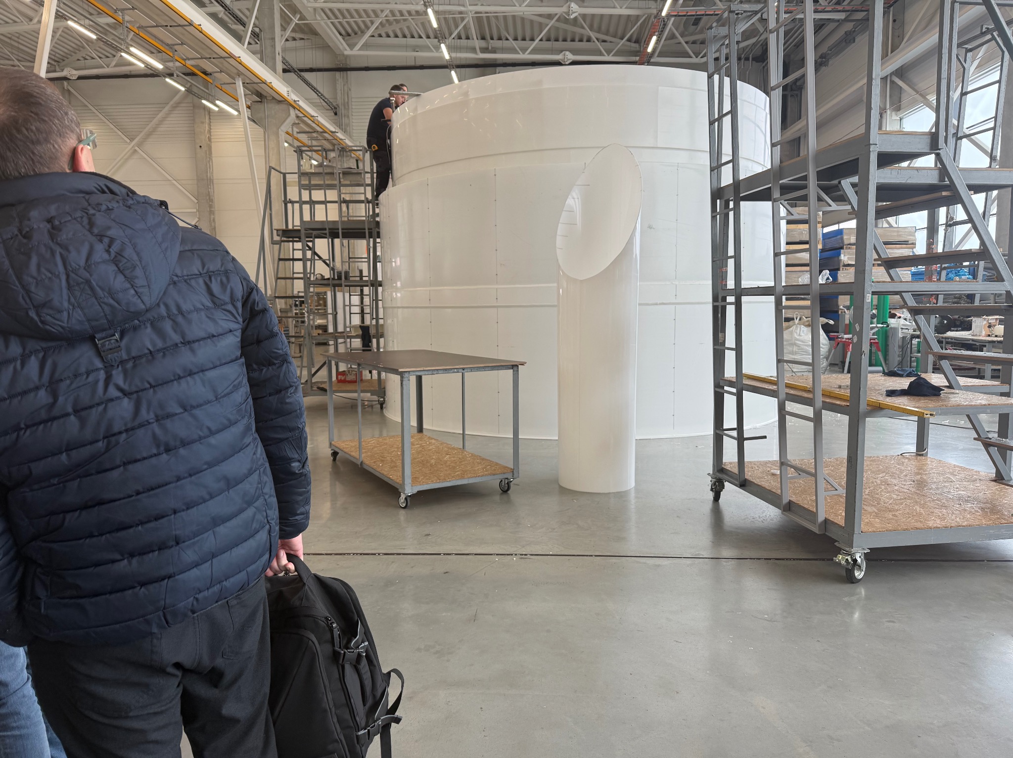 Montaż białego, cylindrycznego zbiornika z rurą boczną w hali produkcyjnej. Pracownik na drabinie, widoczne elementy konstrukcyjne i wózek transportowy.