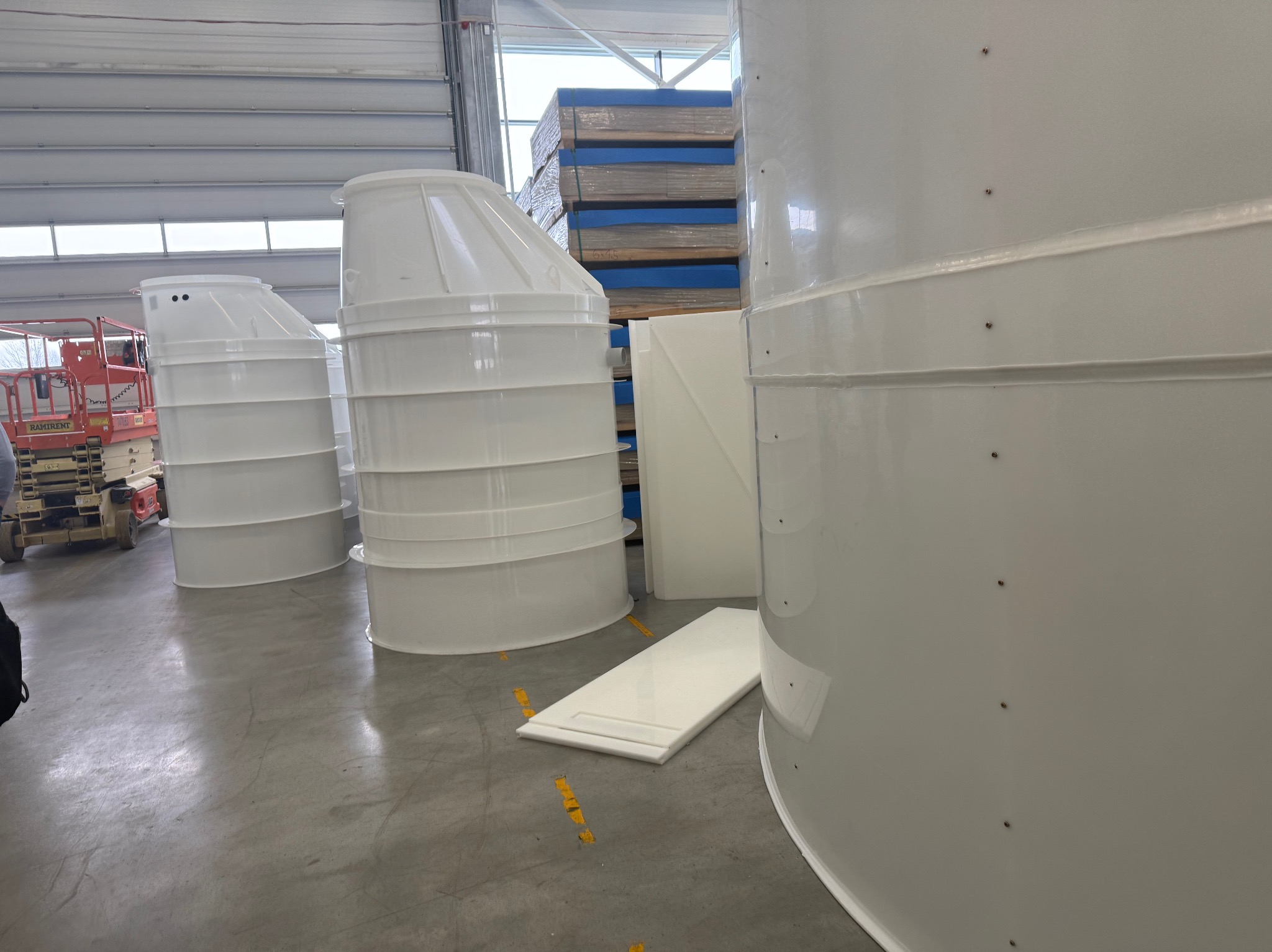 Białe, plastikowe zbiorniki na szambo w hali produkcyjnej, widoczne elementy montażowe i paleta z niebieskim zabezpieczeniem w tle. Lekkie oświetlenie.