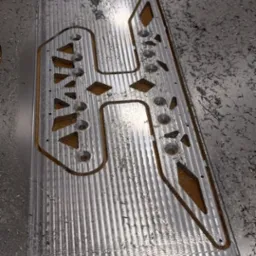 Metalowa płyta po obróbce CNC z wyciętymi wzorami i otworami, otoczona wiórami. Widoczna faktura obrabianej powierzchni.