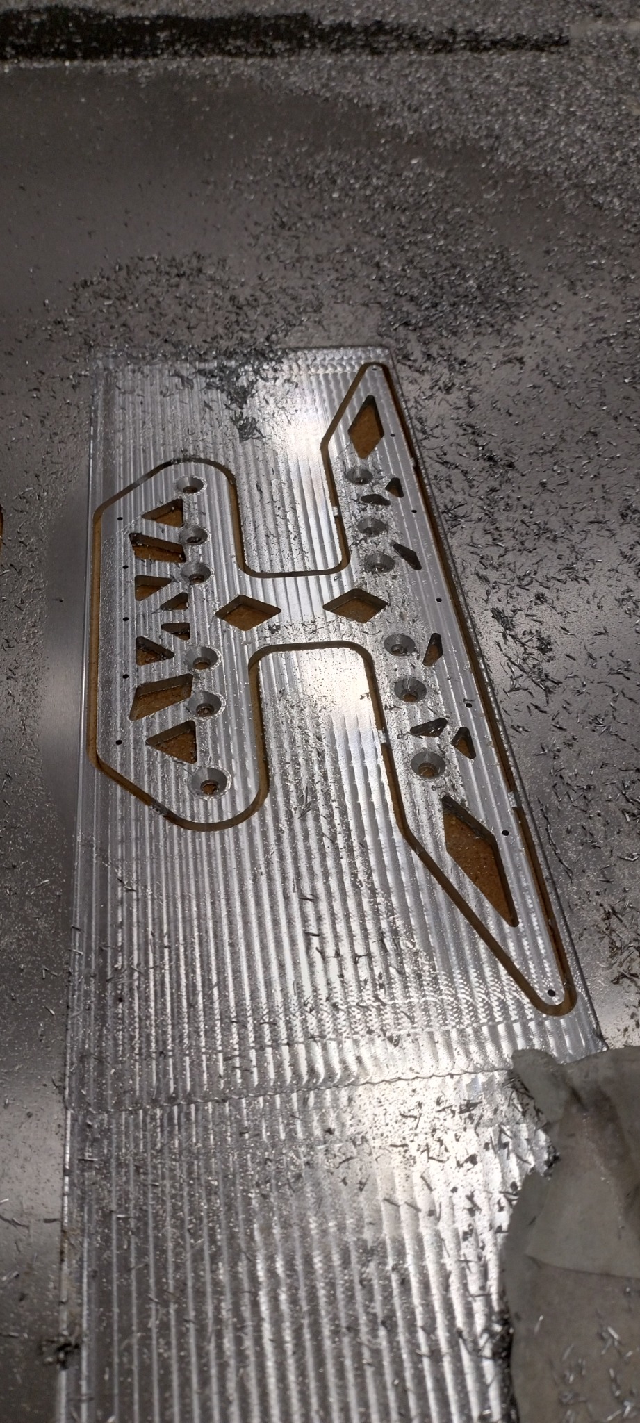 Metalowa płyta po obróbce CNC z wyciętymi wzorami i otworami, otoczona wiórami. Widoczna faktura obrabianej powierzchni.