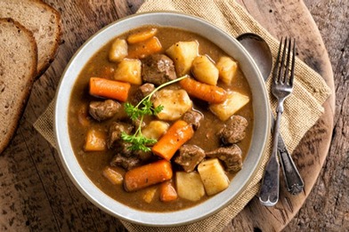 Gulasz wołowy z marchewką i ziemniakami w szarej misce, podany z chlebem na drewnianej desce. Obok widelec i łyżka.