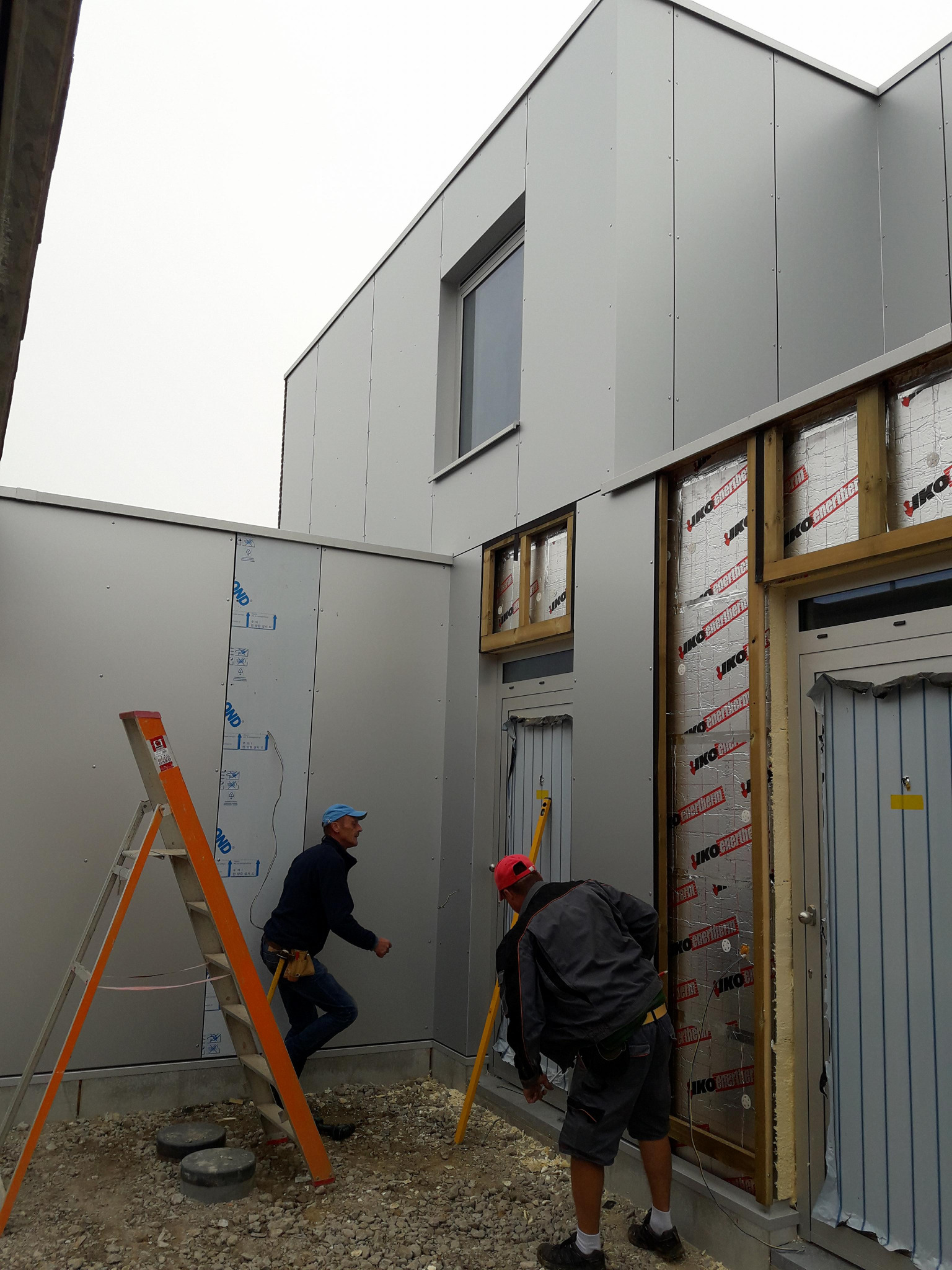 Montaż elewacji budynku z paneli przez dwóch pracowników. Widoczna drabina, izolacja ścian i okna. Prace budowlane w toku, z elementami wykończeniowymi.