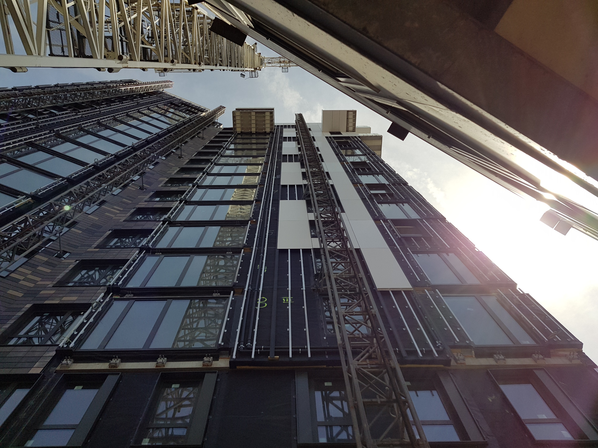 Wysoki budynek w trakcie budowy, widok z dołu. Widoczne okna, rusztowania i fragment żurawia budowlanego. Ujęcie pod dużym kątem.