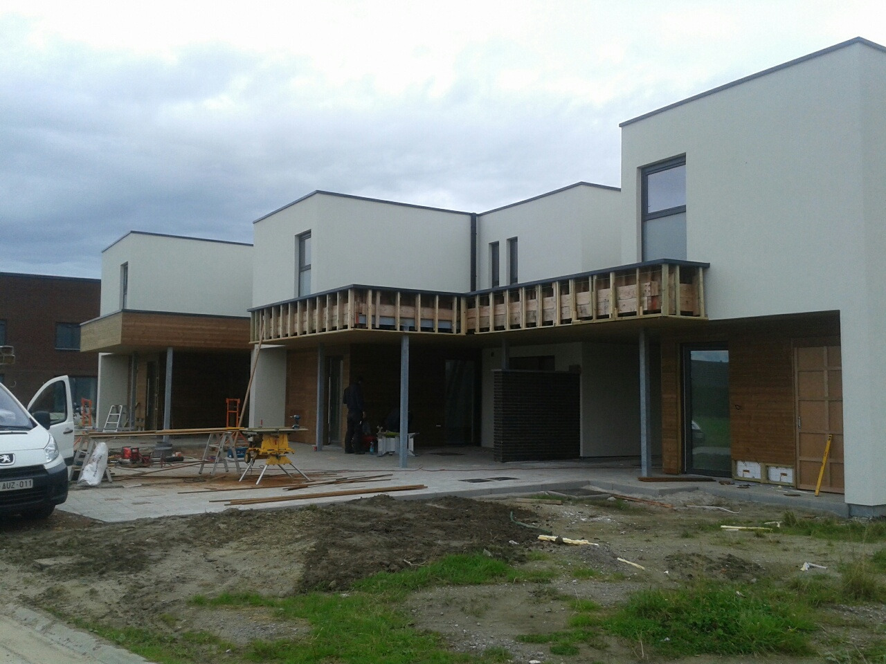 Nowoczesne, parterowe domy w zabudowie szeregowej w trakcie budowy; widoczne drewniane elementy konstrukcyjne balkonów, narzędzia i samochód dostawczy na placu budowy.