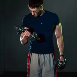 Mężczyzna w okularach ćwiczy biceps z hantlami w mrocznym otoczeniu, oświetlony zielonym światłem. Trening personalny w Krakowie, siłownia.