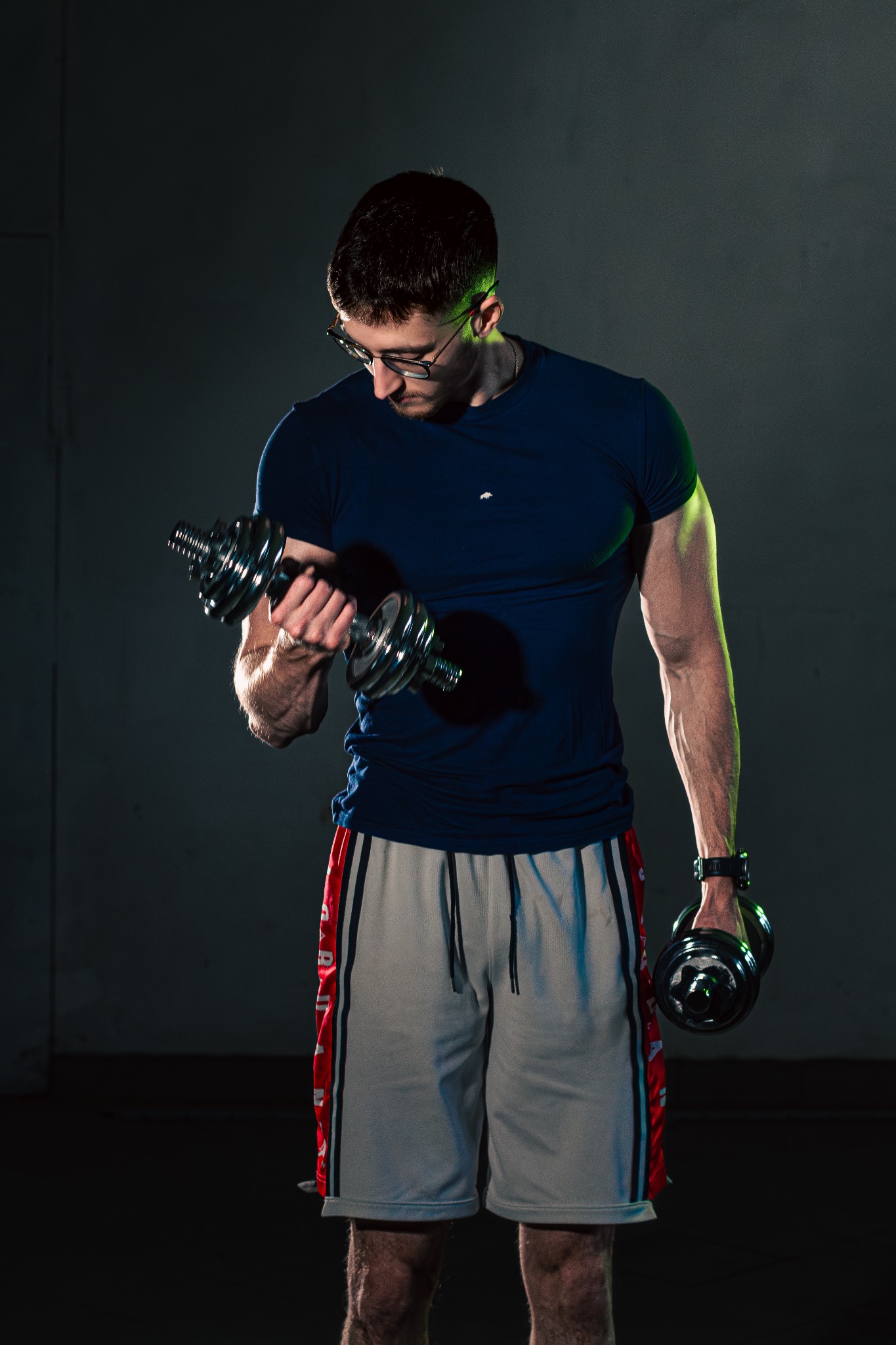 Mężczyzna w okularach ćwiczy biceps z hantlami w mrocznym otoczeniu, oświetlony zielonym światłem. Trening personalny w Krakowie, siłownia.