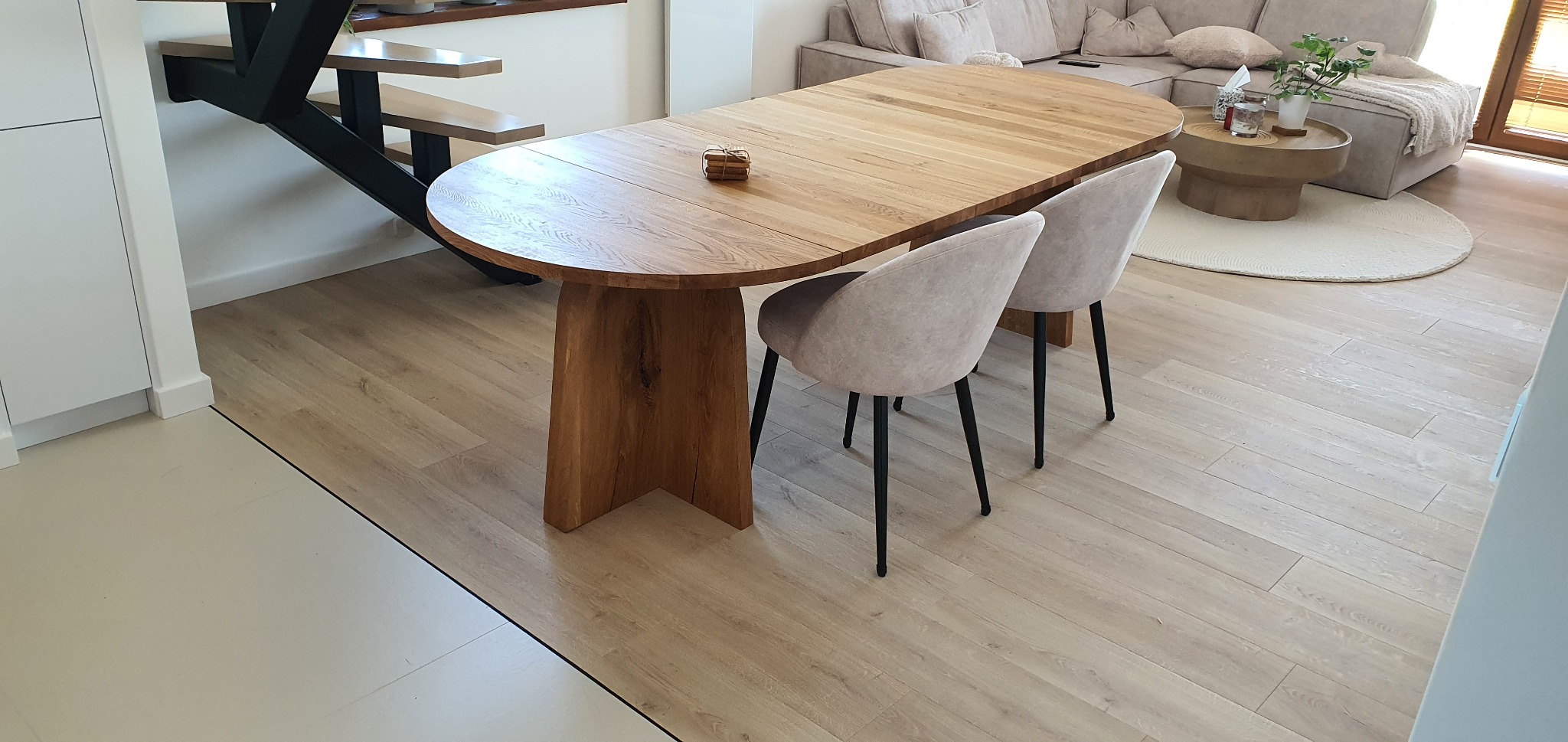 Rozkładany, owalny stół z litego drewna dębowego z dwoma krzesłami w jasnej aranżacji wnętrza, widoczne schody i salon. Naturalne światło podkreśla detale.