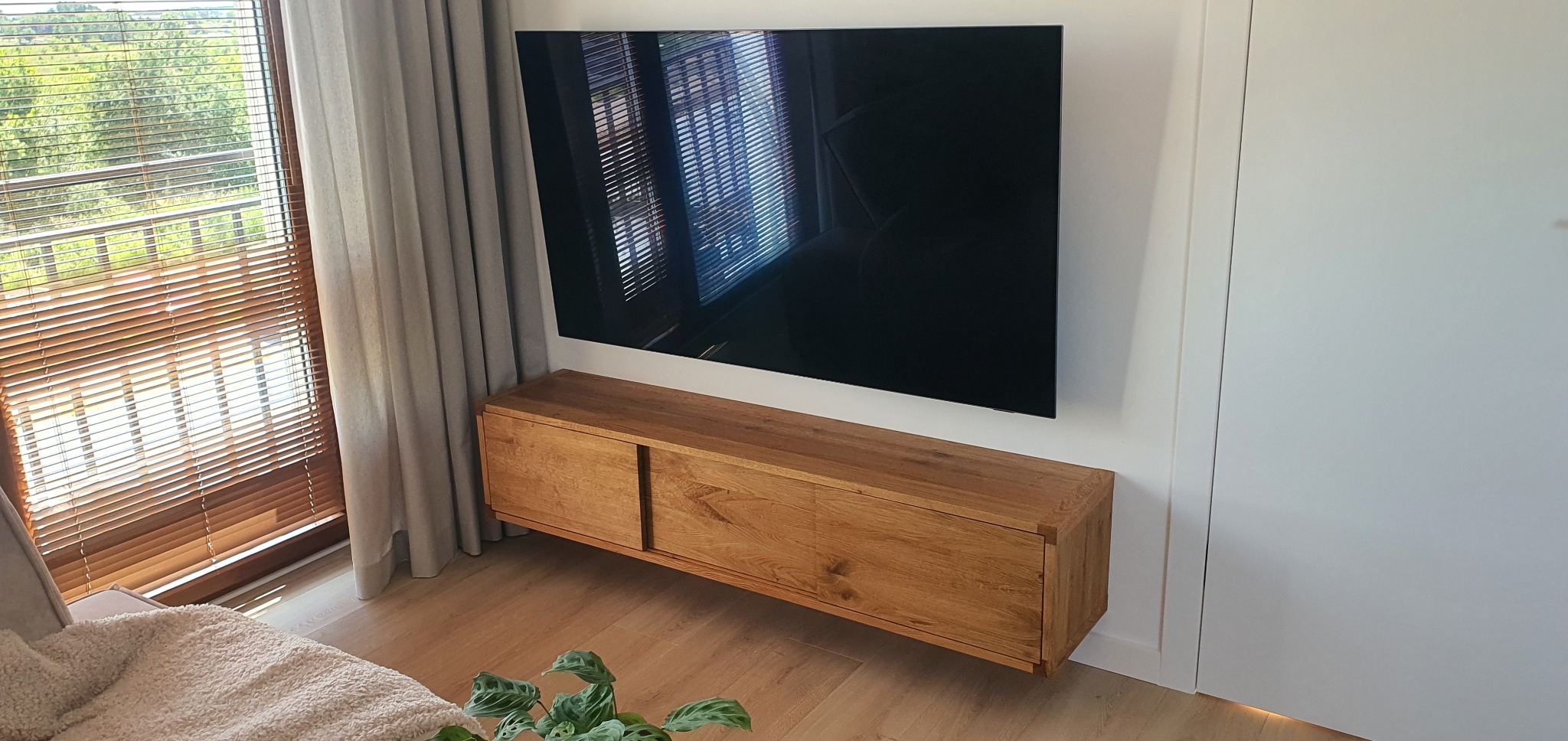 Drewniana szafka RTV z przesuwnymi drzwiami, zamontowana pod telewizorem na ścianie w nowoczesnym salonie z widokiem na zieleń za oknem.