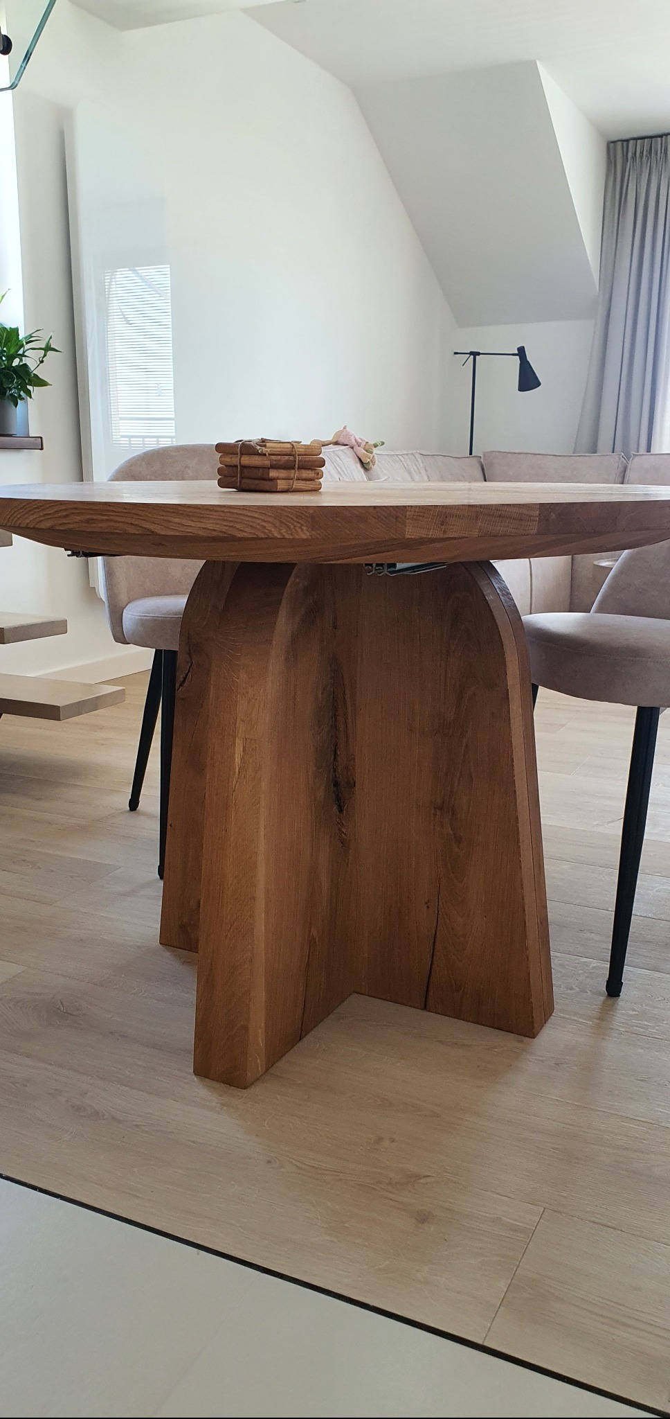 Stół z litego drewna o unikalnej podstawie krzyżowej w jasnym wnętrzu. Na blacie dekoracje, w tle krzesła i sofa w neutralnych barwach.