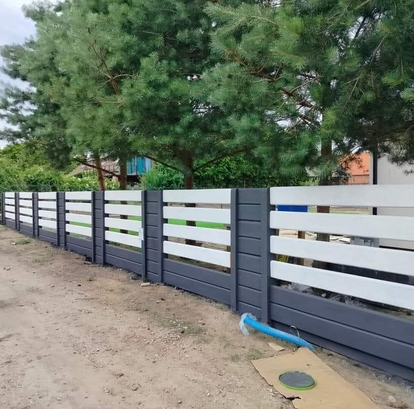 Nowoczesne ogrodzenie betonowe z poziomymi, białymi przęsłami i szarymi słupkami, na tle zieleni drzew. Widoczny fragment niebieskiego węża i kartonu.