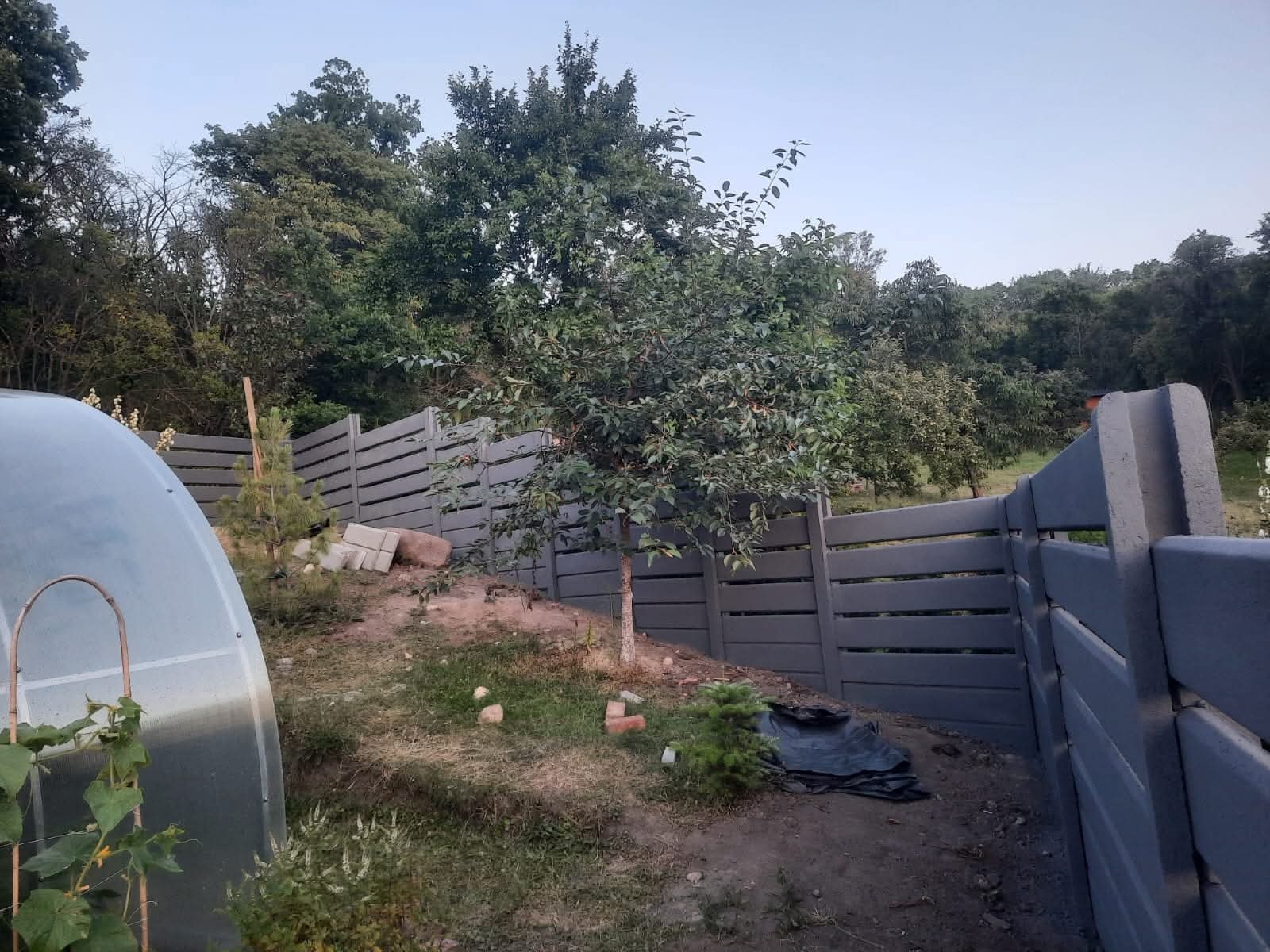 Szary, poziomy płot betonowy z przerwami, otaczający ogród z drzewami i folią. Widoczne elementy konstrukcyjne i nierówny teren.