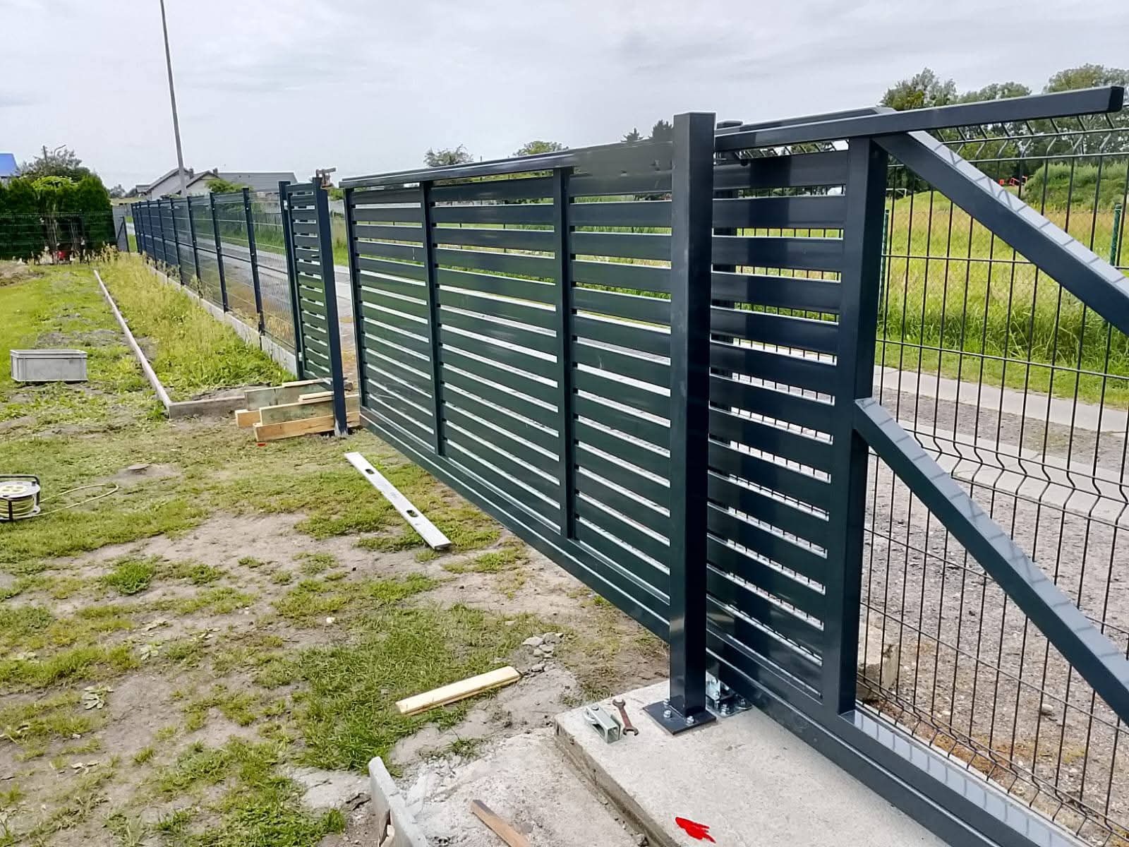 Nowoczesna, przesuwna brama metalowa z poziomymi panelami, zamontowana na betonowym fundamencie. W tle widoczne ogrodzenie panelowe i trawnik.