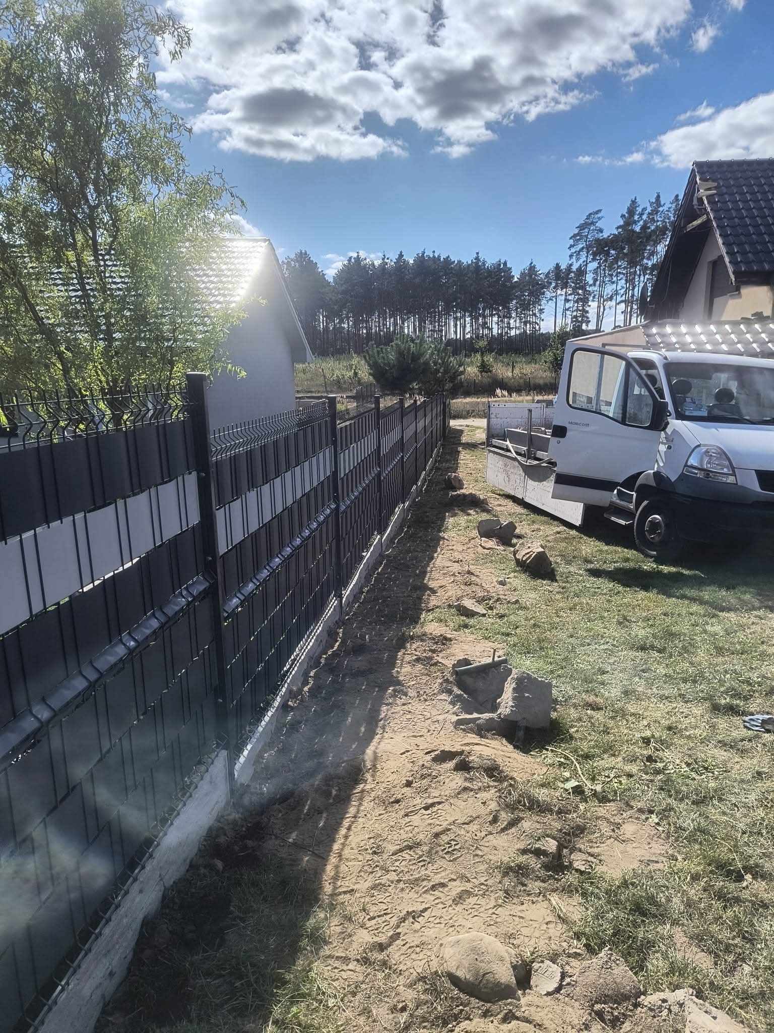 Nowoczesne ogrodzenie panelowe z szarymi wypełnieniami, świeżo zamontowane na betonowej podmurówce, z widocznym samochodem dostawczym w tle.