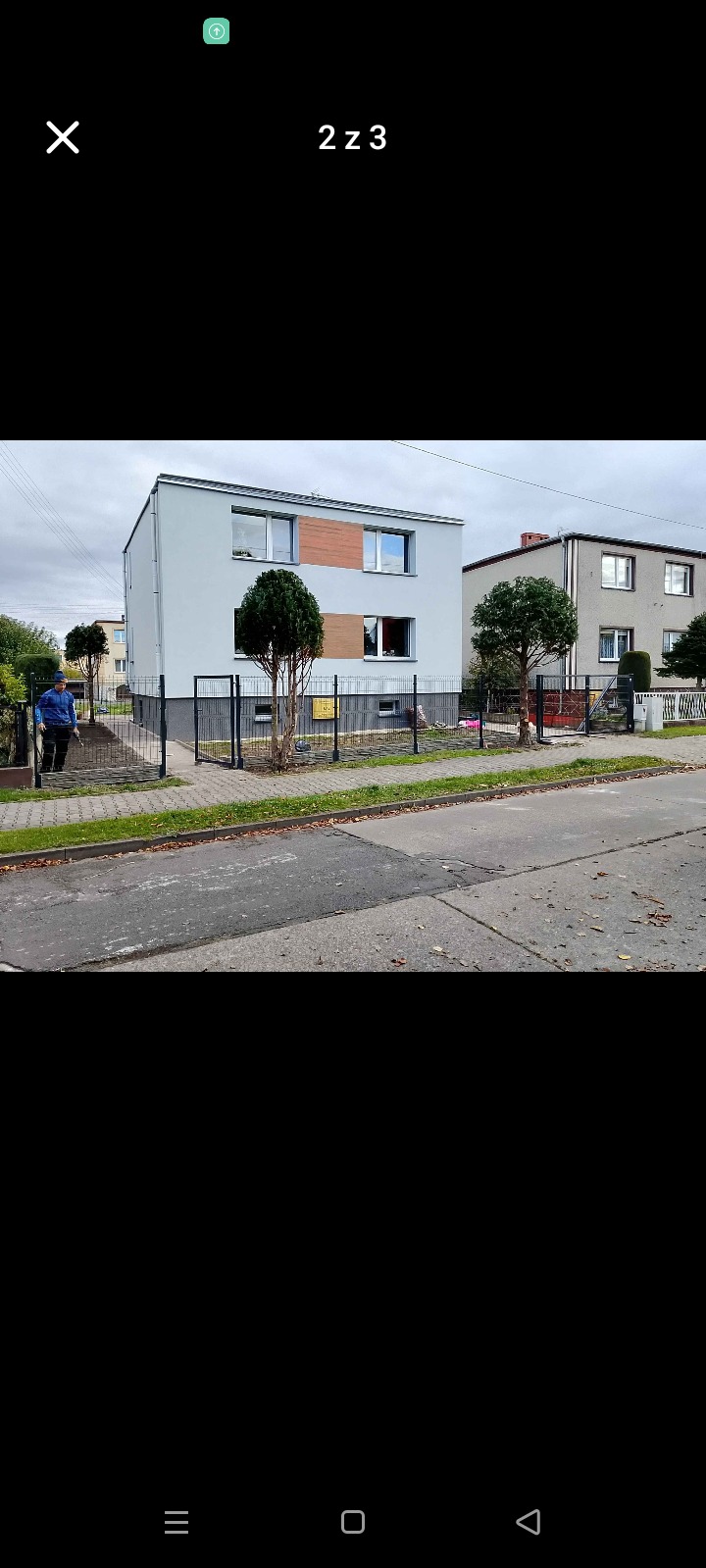 Nowoczesny dom z szarą elewacją i czarnym ogrodzeniem panelowym. Osoba w niebieskiej kurtce wchodzi przez furtkę. Dwa małe drzewka przed budynkiem.