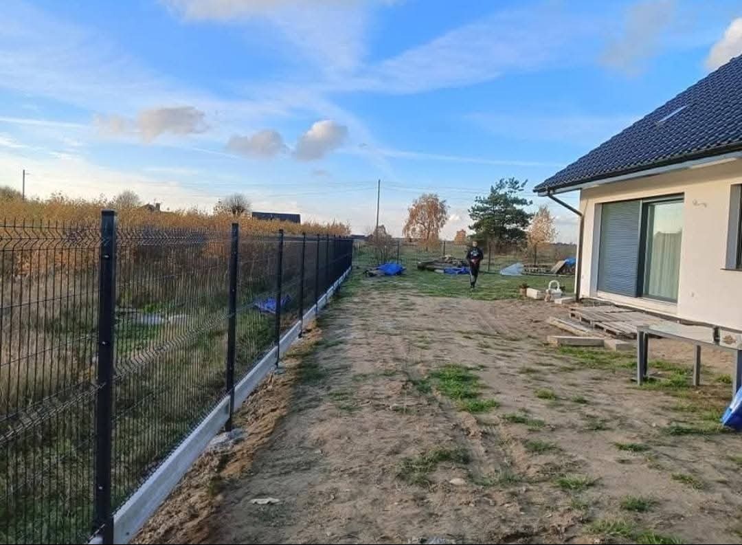 Nowoczesne ogrodzenie panelowe z podmurówką betonową, widoczne na tle domu jednorodzinnego i błękitnego nieba. Osoba w oddali dodaje skali.