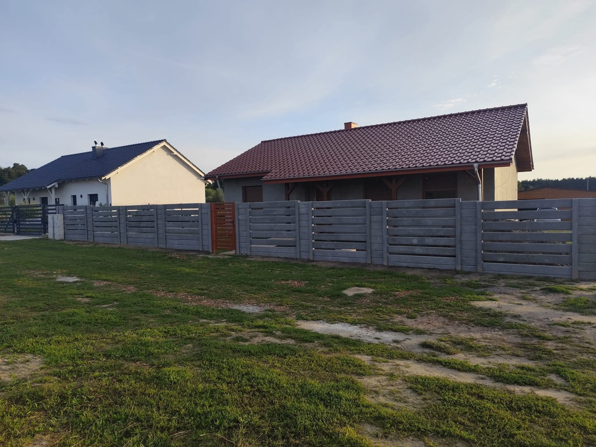 Szary, poziomy płot betonowy z drewnianą bramką, oddzielający posesje z domami o różnej architekturze, na tle zielonej trawy i błękitnego nieba.