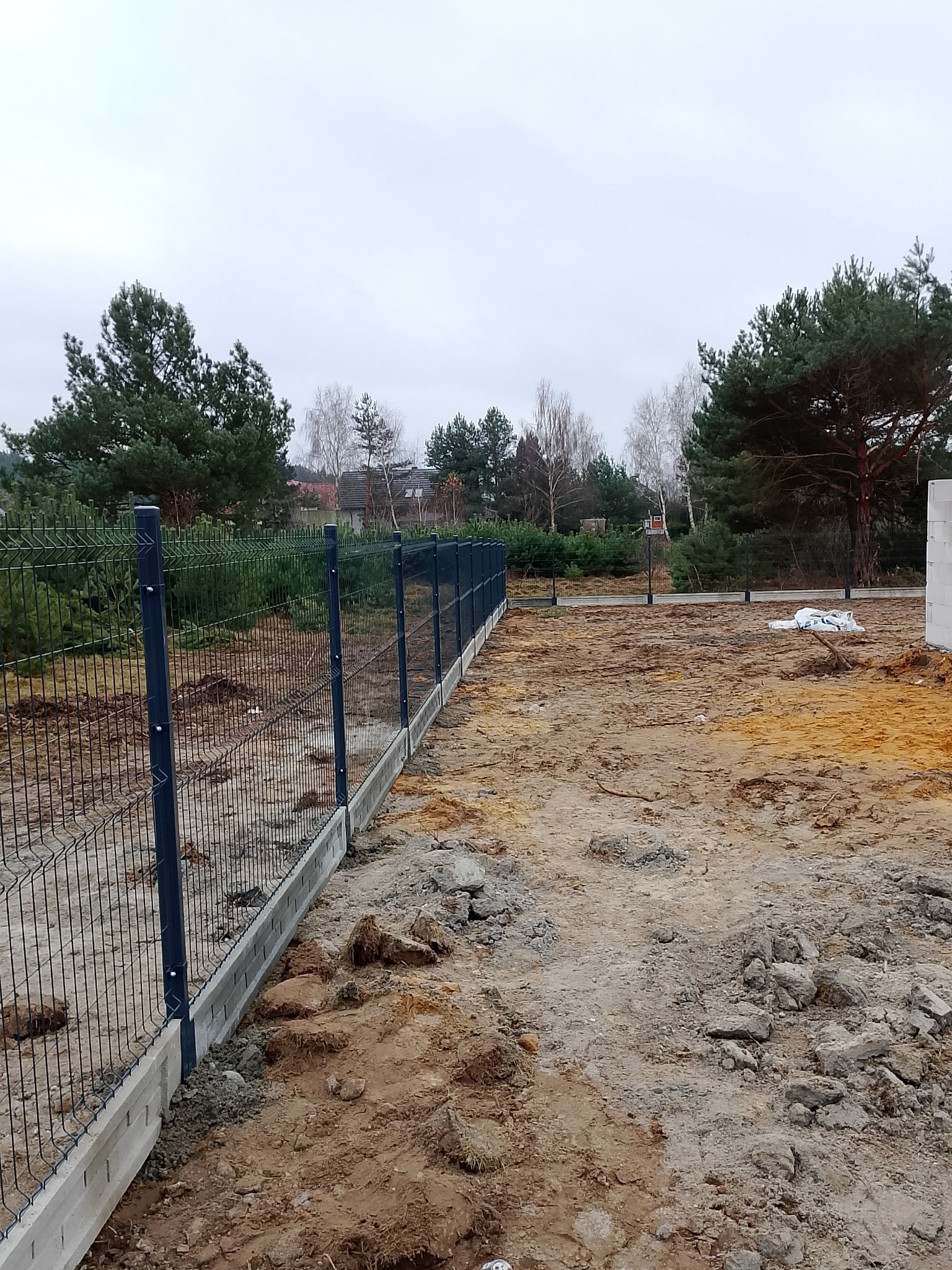 Nowoczesne ogrodzenie panelowe z podmurówką betonową na działce w trakcie budowy, widok z perspektywy ziemi, pochmurne niebo w tle.