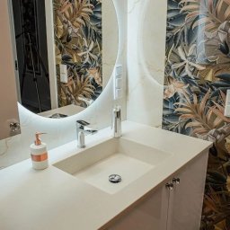 Mateusz Groszek - Nowoczesna łazienka z umywalką wpuszczaną w blat, okrągłym lustrem z podświetleniem LED i tapetą w tropikalne liście. Bateria chromowana i dozownik mydła dodają elegancji.