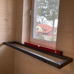 Mateusz Groszek - Metalowa rama pod oknem, poziomica na górze, wnętrze w płytkach. Widok z okna na domy i drzewa. Konstrukcja w trakcie montażu, surowy wygląd metalu.