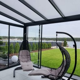 Mateusz Groszek - Nowoczesna zabudowa balkonu z czarnymi ramami i szklanymi ścianami, widok na jezioro i ogród. Dwa wiszące fotele z poduszkami wewnątrz.