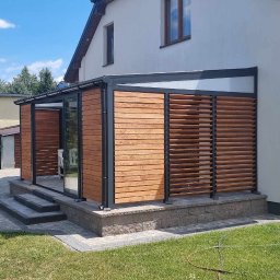 Mateusz Groszek - Nowoczesna zabudowa balkonu z drewnianymi lamelami i szklaną ścianą, widoczna z zewnątrz, na tle domu z jasną elewacją i zielonego trawnika.