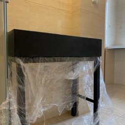 Mateusz Groszek - Czarna szafka łazienkowa z metalowym stelażem, zabezpieczona folią ochronną. Jasne płytki na ścianach i podłodze. Widoczny fragment kabiny prysznicowej.