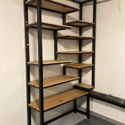 Mateusz Groszek - Ręcznie robiony regał z czarnego metalu i drewna dębowego, wkomponowany w narożnik pomieszczenia. Półki o nieregularnym kształcie tworzą unikalny design.