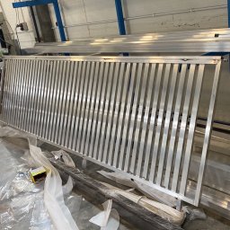 Mateusz Groszek - Surowa, aluminiowa konstrukcja balustrady z pionowymi prętami w warsztacie. W tle maszyny i narzędzia ślusarskie, elementy konstrukcyjne. Widoczne ślady obróbki.