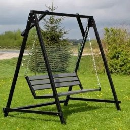 Mateusz Groszek - Metalowa huśtawka ogrodowa z siedziskiem z desek, zawieszona na łańcuchach, na tle zielonego trawnika i krzewów, z widocznym fragmentem drogi za ogrodzeniem.