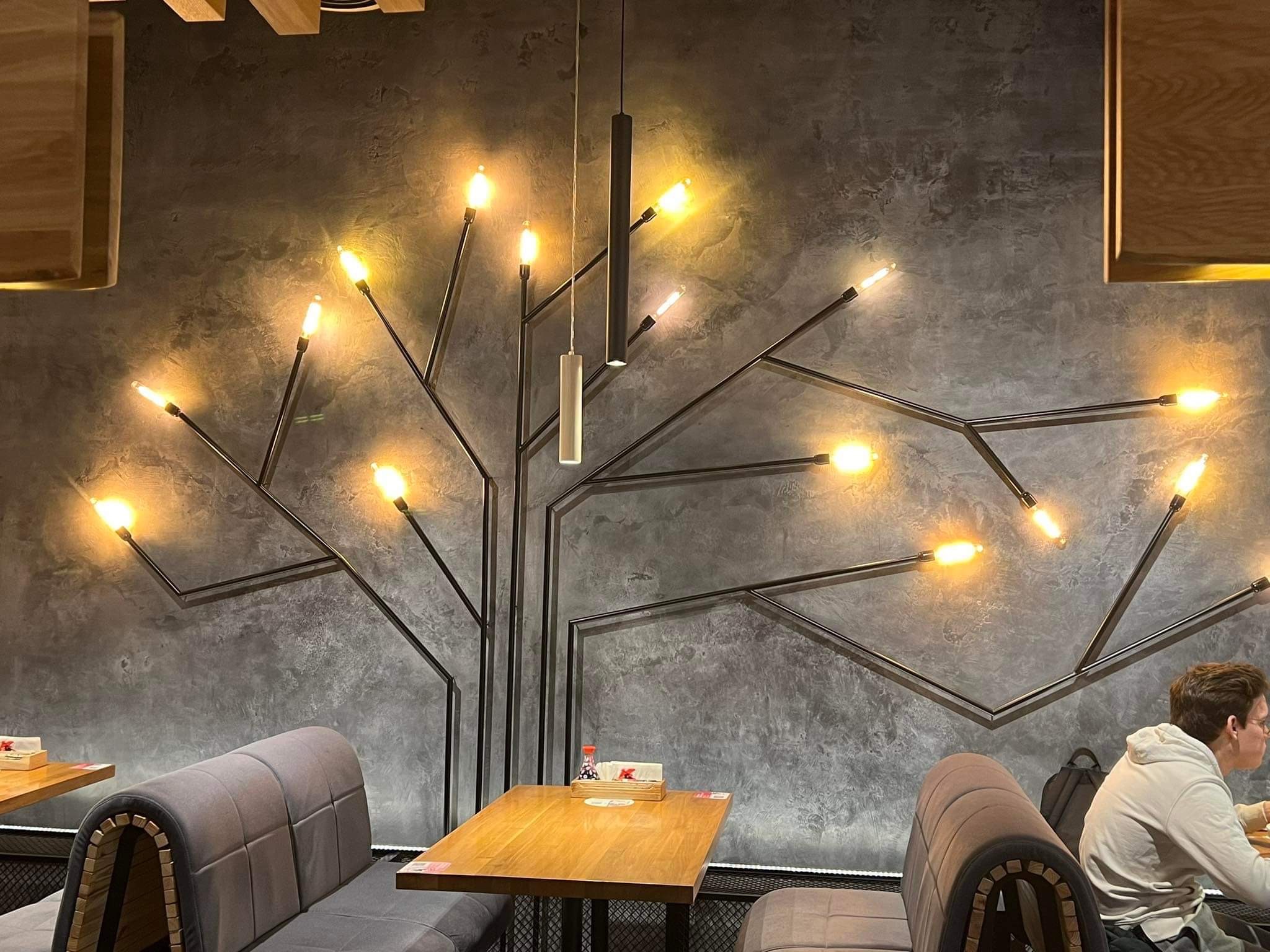 Metalowa konstrukcja przypominająca drzewo z żarówkami na szarym tle. Element dekoracyjny w restauracji lub kawiarni, oświetlający przestrzeń. Widoczny stolik i krzesła.