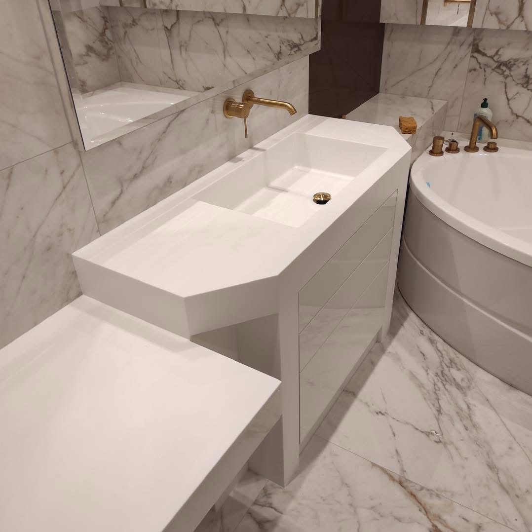 Nowoczesna, biała umywalka z szafką i blatem w łazience wykończonej marmurem, złota armatura, okrągła wanna. Minimalistyczny design.