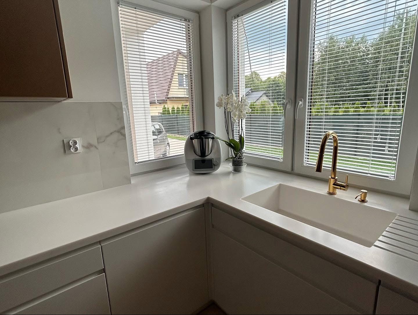 Biały, minimalistyczny blat kuchenny z oknem i złotą armaturą. Widoczne rolety, kwiaty i sprzęt kuchenny. Ujęcie z bliska, skupione na detalu i estetyce.