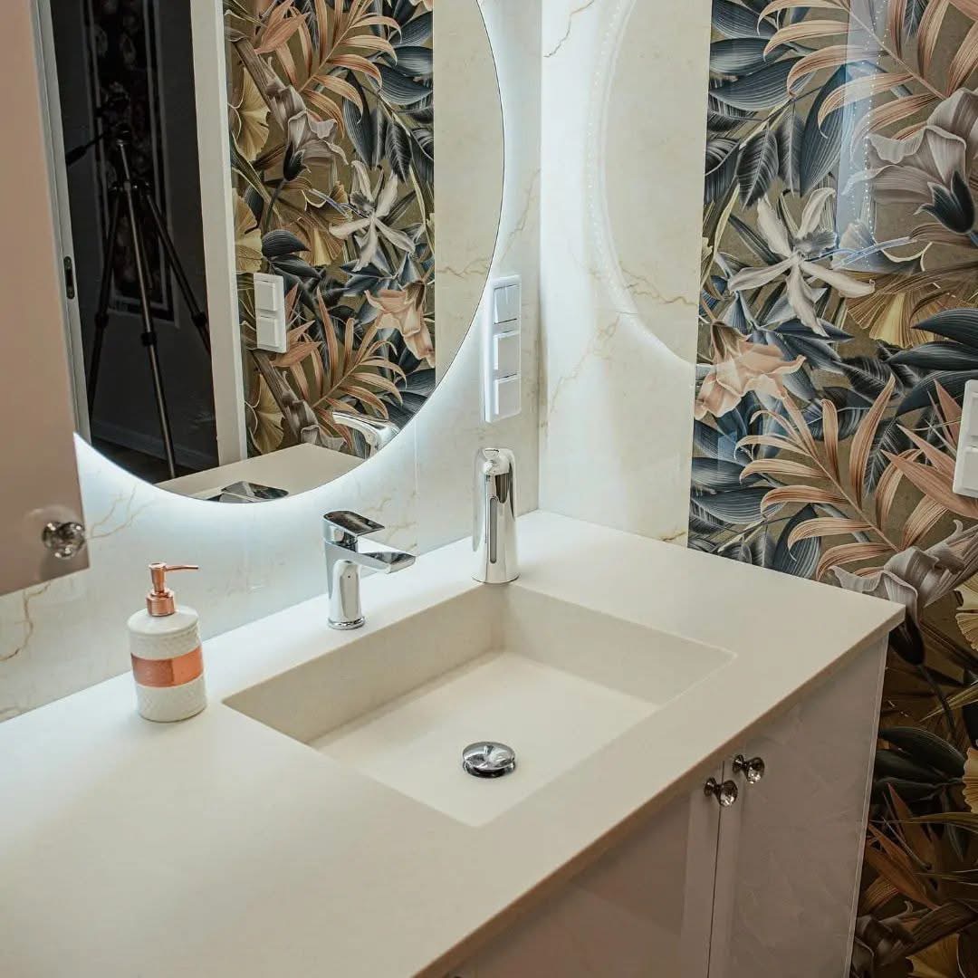 Nowoczesna łazienka z umywalką wpuszczaną w blat, okrągłym lustrem z podświetleniem LED i tapetą w tropikalne liście. Bateria chromowana i dozownik mydła dodają elegancji.