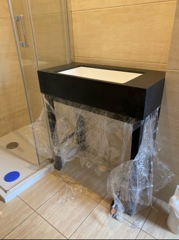 Nowoczesna, czarna szafka łazienkowa z białą umywalką, zabezpieczona folią ochronną, stoi obok kabiny prysznicowej w jasnej łazience.