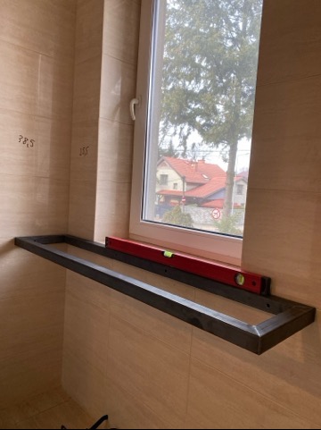 Metalowa rama pod oknem, poziomica na górze, wnętrze w płytkach. Widok z okna na domy i drzewa. Konstrukcja w trakcie montażu, surowy wygląd metalu.