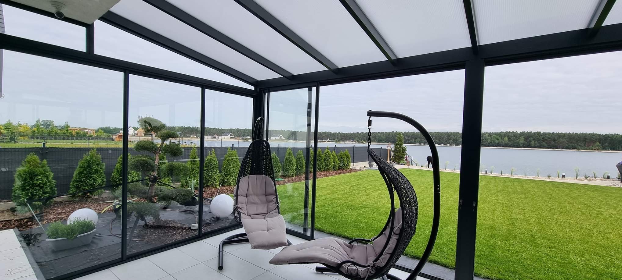 Nowoczesna zabudowa balkonu z czarnymi ramami i szklanymi ścianami, widok na jezioro i ogród. Dwa wiszące fotele z poduszkami wewnątrz.