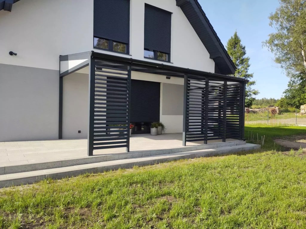 Nowoczesna, czarna pergola metalowa z poziomymi lamelami, przylegająca do białego domu z szarym akcentem. Widok z zewnątrz, trawnik na pierwszym planie.