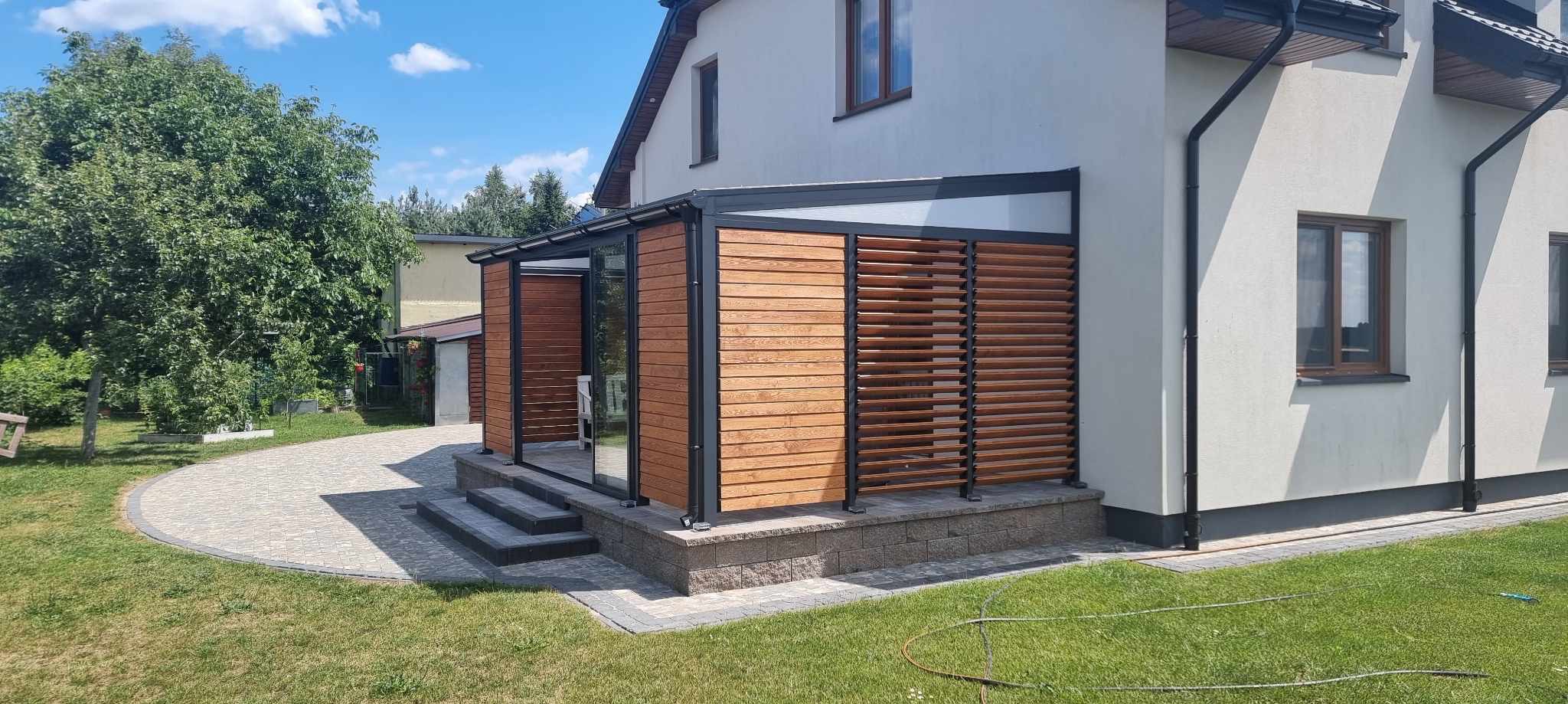 Nowoczesna zabudowa balkonu z drewnianymi lamelami i szklaną ścianą, widoczna z zewnątrz, na tle domu z jasną elewacją i zielonego trawnika.
