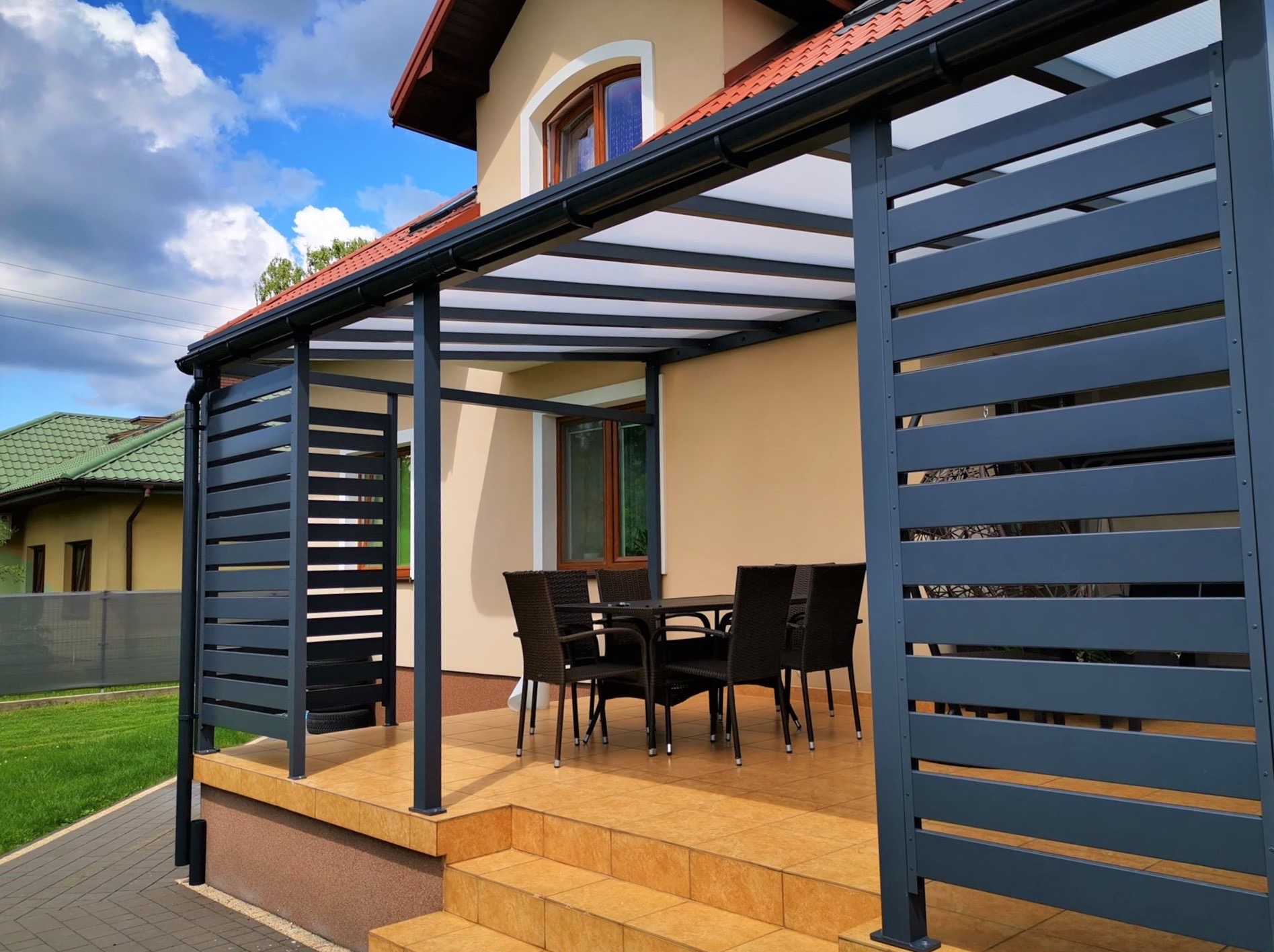 Nowoczesna pergola z metalową konstrukcją i ażurowymi ścianami, tworząca zacienioną przestrzeń na tarasie z meblami ogrodowymi. Widok na dom i błękitne niebo.