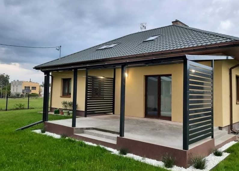 Nowoczesna pergola z metalowymi panelami, tworząca zacienioną przestrzeń tarasową przy domu z zielonym dachem i ozdobnym obramowaniem z białych kamieni.