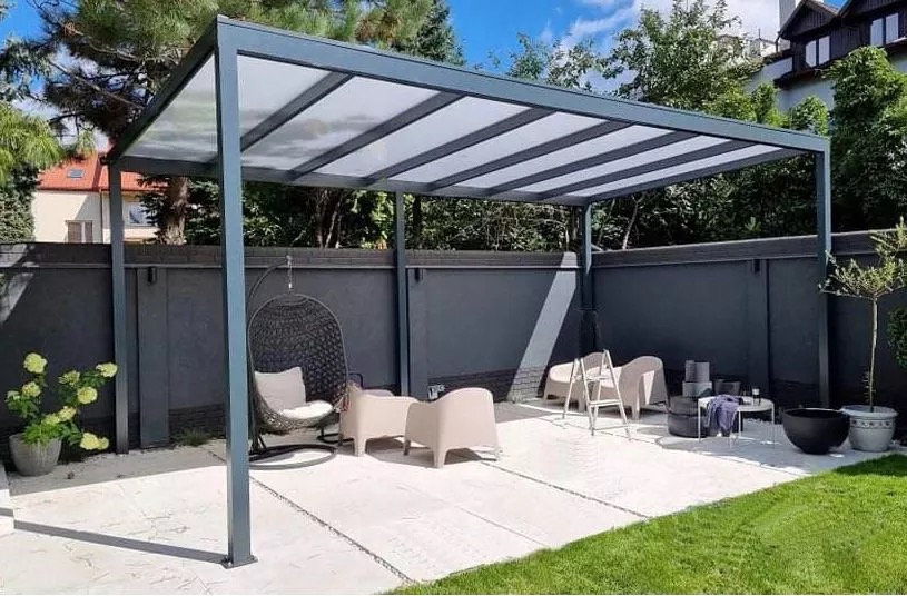 Nowoczesna pergola z szarą metalową konstrukcją i przezroczystym zadaszeniem, otoczona zielenią ogrodu, z meblami wypoczynkowymi. Widok z niskiego kąta.