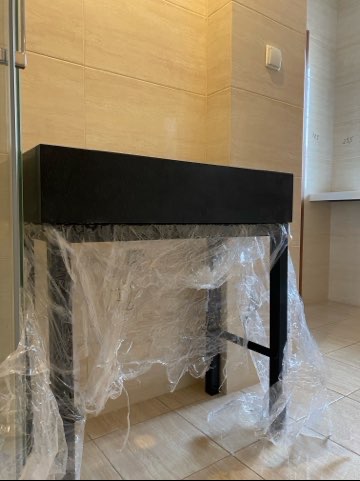 Czarna szafka łazienkowa z metalowym stelażem, zabezpieczona folią ochronną. Jasne płytki na ścianach i podłodze. Widoczny fragment kabiny prysznicowej.