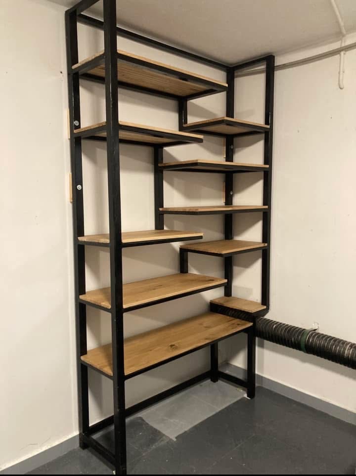 Ręcznie robiony regał z czarnego metalu i drewna dębowego, wkomponowany w narożnik pomieszczenia. Półki o nieregularnym kształcie tworzą unikalny design.