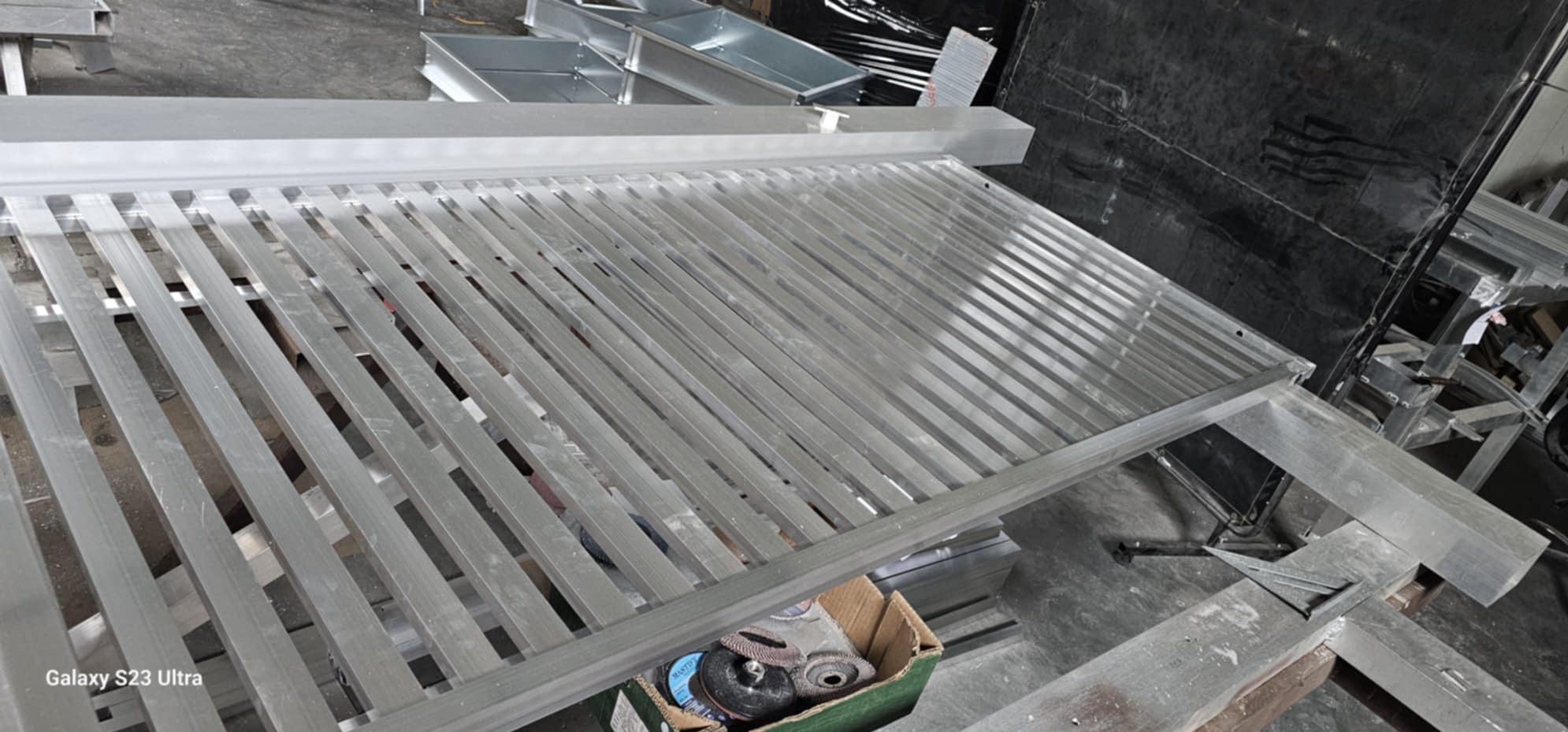 Metalowa konstrukcja balustrady z profili, widoczne narzędzia i elementy montażowe w tle. Szlifowany metal w surowym stanie, przygotowany do dalszej obróbki.