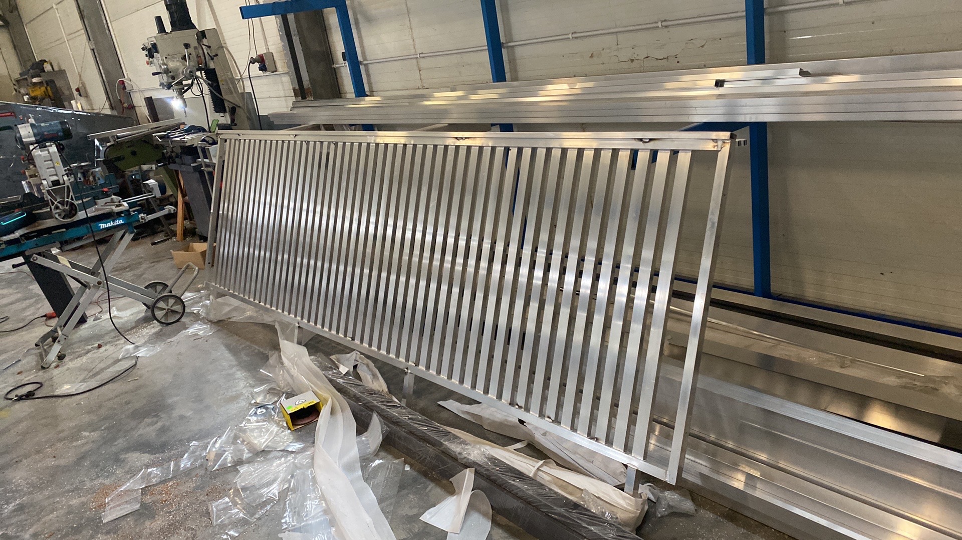 Surowa, aluminiowa konstrukcja balustrady z pionowymi prętami w warsztacie. W tle maszyny i narzędzia ślusarskie, elementy konstrukcyjne. Widoczne ślady obróbki.
