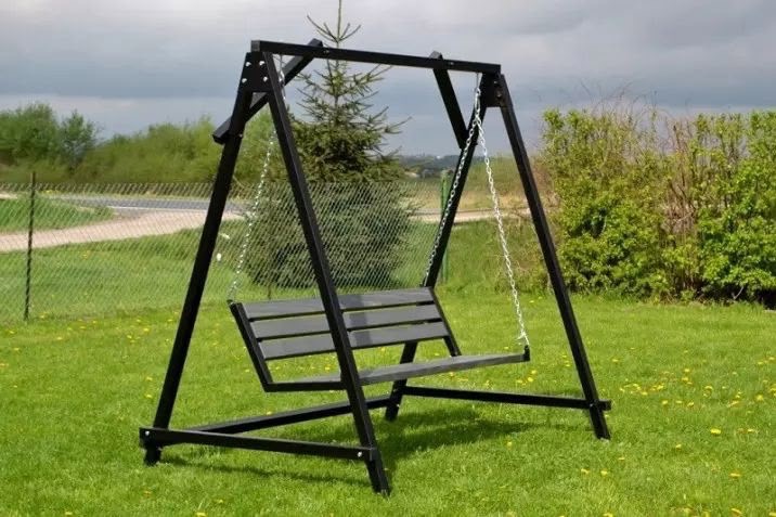 Metalowa huśtawka ogrodowa z siedziskiem z desek, zawieszona na łańcuchach, na tle zielonego trawnika i krzewów, z widocznym fragmentem drogi za ogrodzeniem.