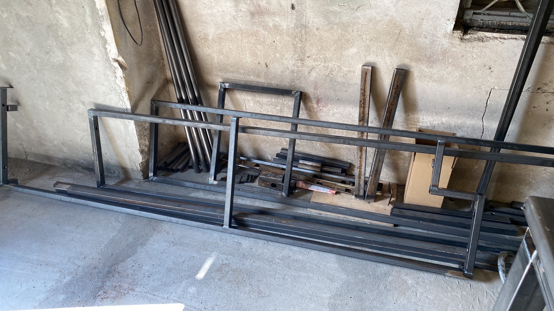 Elementy konstrukcyjne z metalu, prawdopodobnie w trakcie produkcji, ułożone na podłodze w warsztacie. Widoczne spawane ramy i profile, w tle surowa ściana.
