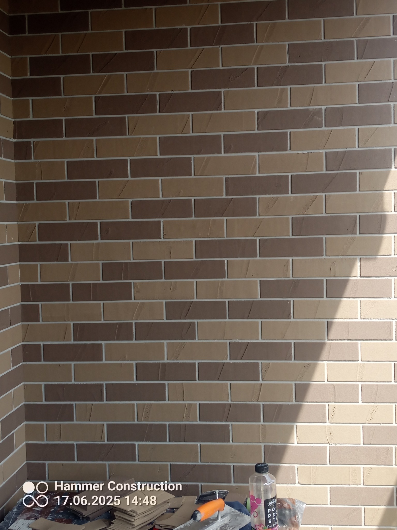 Dekoracyjna ściana z cegieł klinkierowych w odcieniach beżu i brązu, z widocznym cieniem i fragmentem narzędzi na dole kadru. Nowoczesna elewacja.