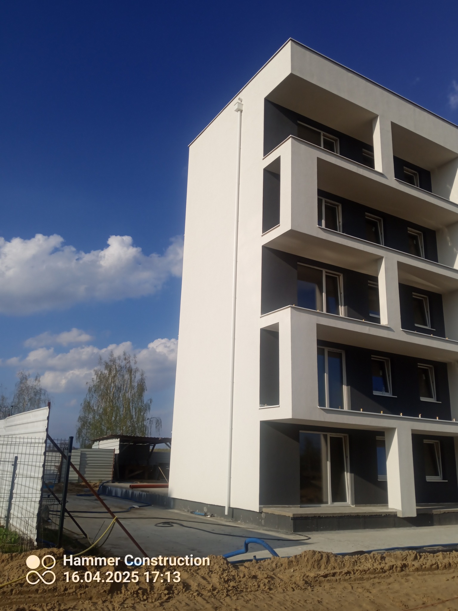 Nowoczesny, wielopiętrowy budynek z białymi elementami elewacji i ciemnymi balkonami, na tle błękitnego nieba z chmurami. Widoczne prace wykończeniowe wokół budynku.